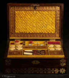 SB524: Rosewood and brass inlaid sewing box in high Regency style circa 1815. A box veneered in beautifully figured rosewood and inlaid with brass. The design of the inlay is of highly stylized flora, suggesting neoclassical designs hinting at trefoil motifs. The juxtaposition of dark wood with bold brass inlay was popular in the early part of the 19th century. The wood and the bright brass were mutually enhancing. The box retains its original  fully fitted tray covered in yellow paper with gold embossed supplementary lids. There is a set of  eight turned and carved vegetable ivory  spools. Enlarge Picture